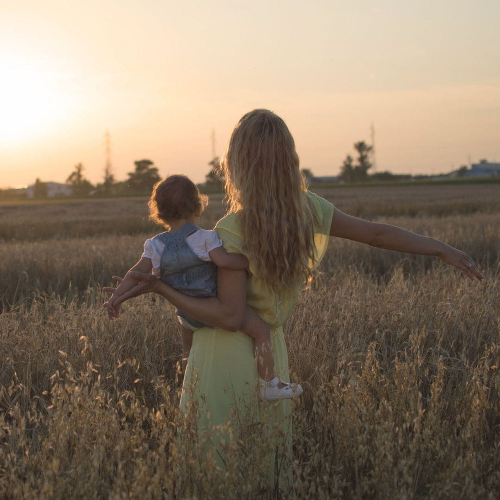 Mother carrying a young child through a golden meadow at sunset, reflecting Edenwild’s family-rooted ancestral skincare philosophy.