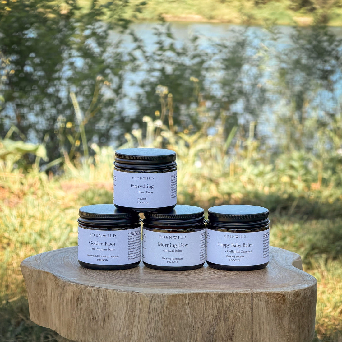 Four Edenwild tallow balms displayed on a wood stump outdoors — Everything Balm, Golden Root, Morning Dew, and Happy Baby Balm