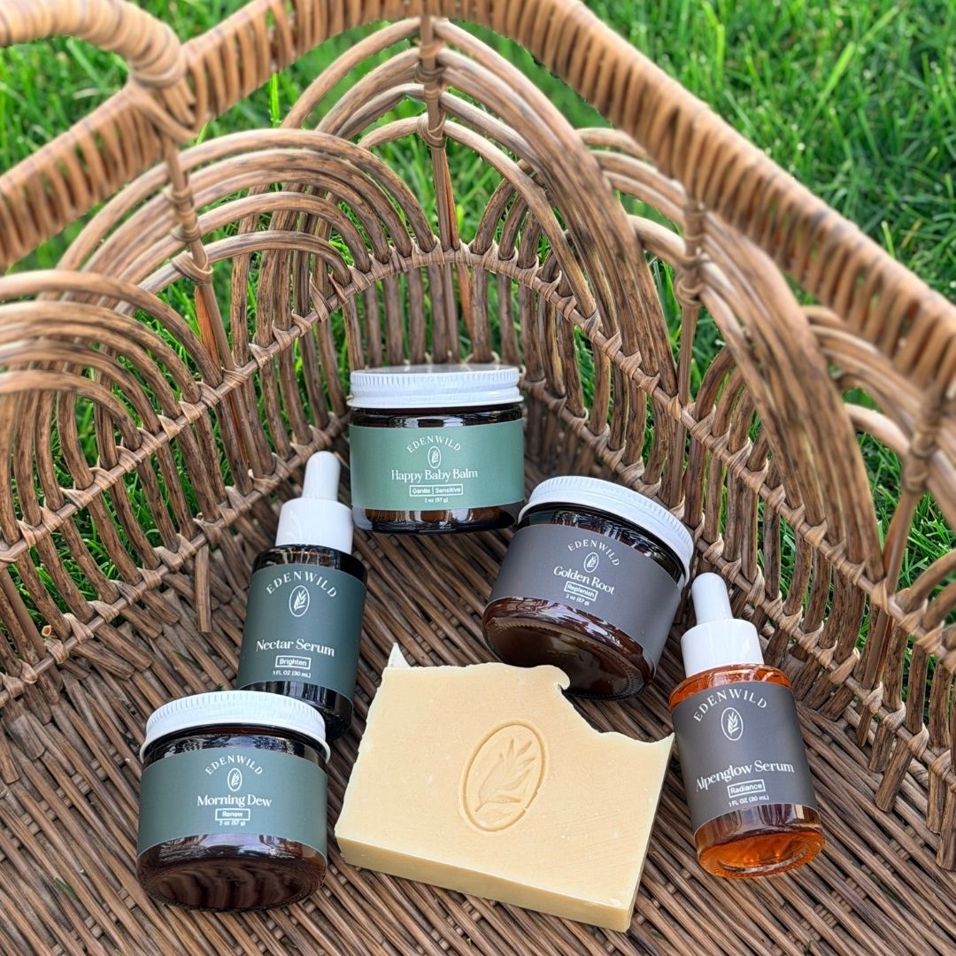 denwild ancestral skincare products arranged in a woven basket on grass, including tallow balms, facial serums, and a soap bar.