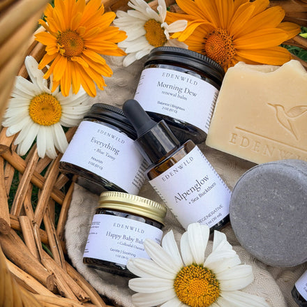 Natural skincare routine products by Edenwild — Everything Balm, Morning Dew renewal balm, Alpenglow sea buckthorn serum, and Happy Baby Balm arranged in a wicker basket with fresh daisies and marigolds