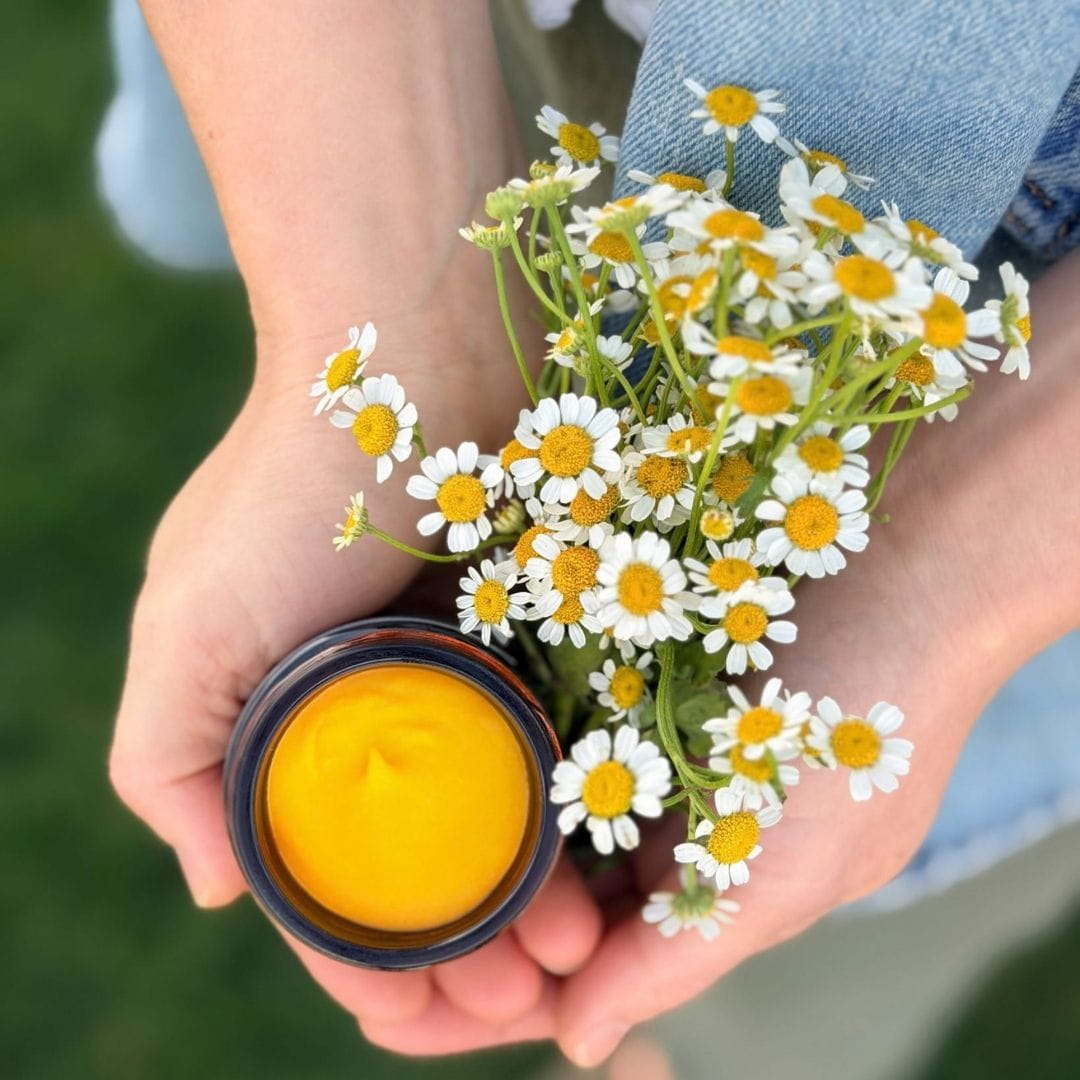 I’ll keep it descriptive and weave in your ancestral skincare positioning naturally. Hands holding an open jar of golden skincare balm beside white chamomile flowers, reflecting Edenwild’s botanical approach to ancestral skincare.