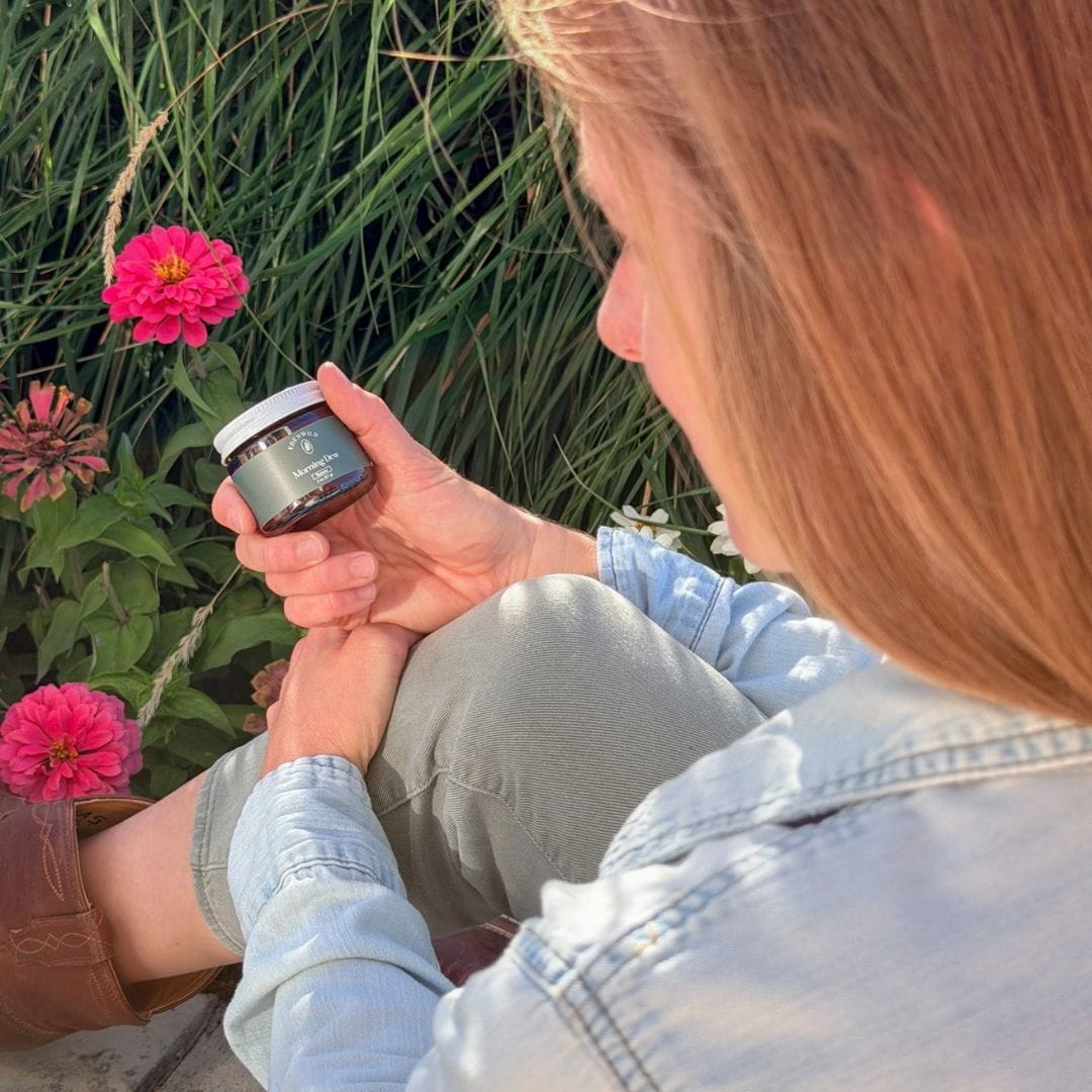 Woman holding an Edenwild skincare balm jar beside pink garden flowers, reflecting the brand’s ancestral skincare roots.