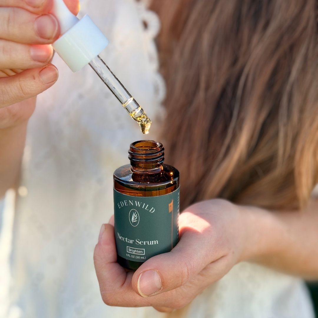 Hands holding Edenwild Nectar Serum in an amber glass bottle, with a dropper releasing a golden drop of brightening facial serum.
