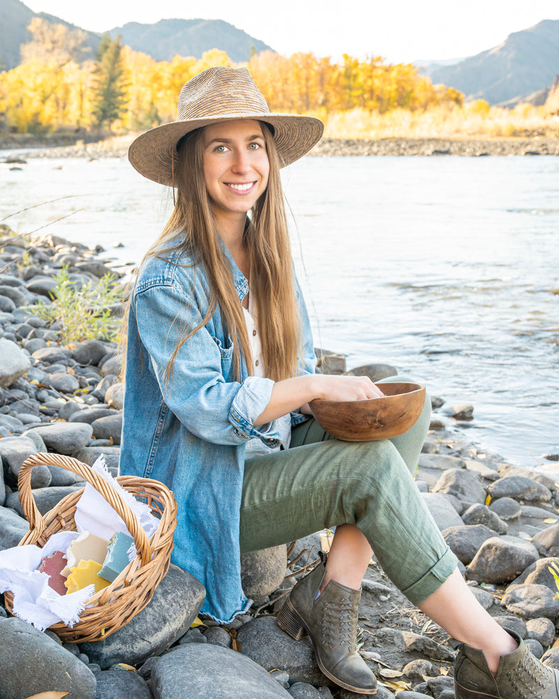 Angela Clifton, founder of Edenwild natural skincare, sitting on a Wyoming riverbank with a basket of botanical products