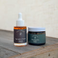 Edenwild Alpenglow Serum dropper bottle and Morning Dew balm jar on a rustic wooden surface, paired as an ancestral skincare routine.