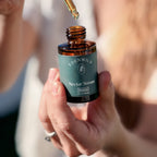Close-up of a hand holding Edenwild Nectar Serum in an amber glass bottle, with a dropper of golden facial oil above the opening in sunlight.