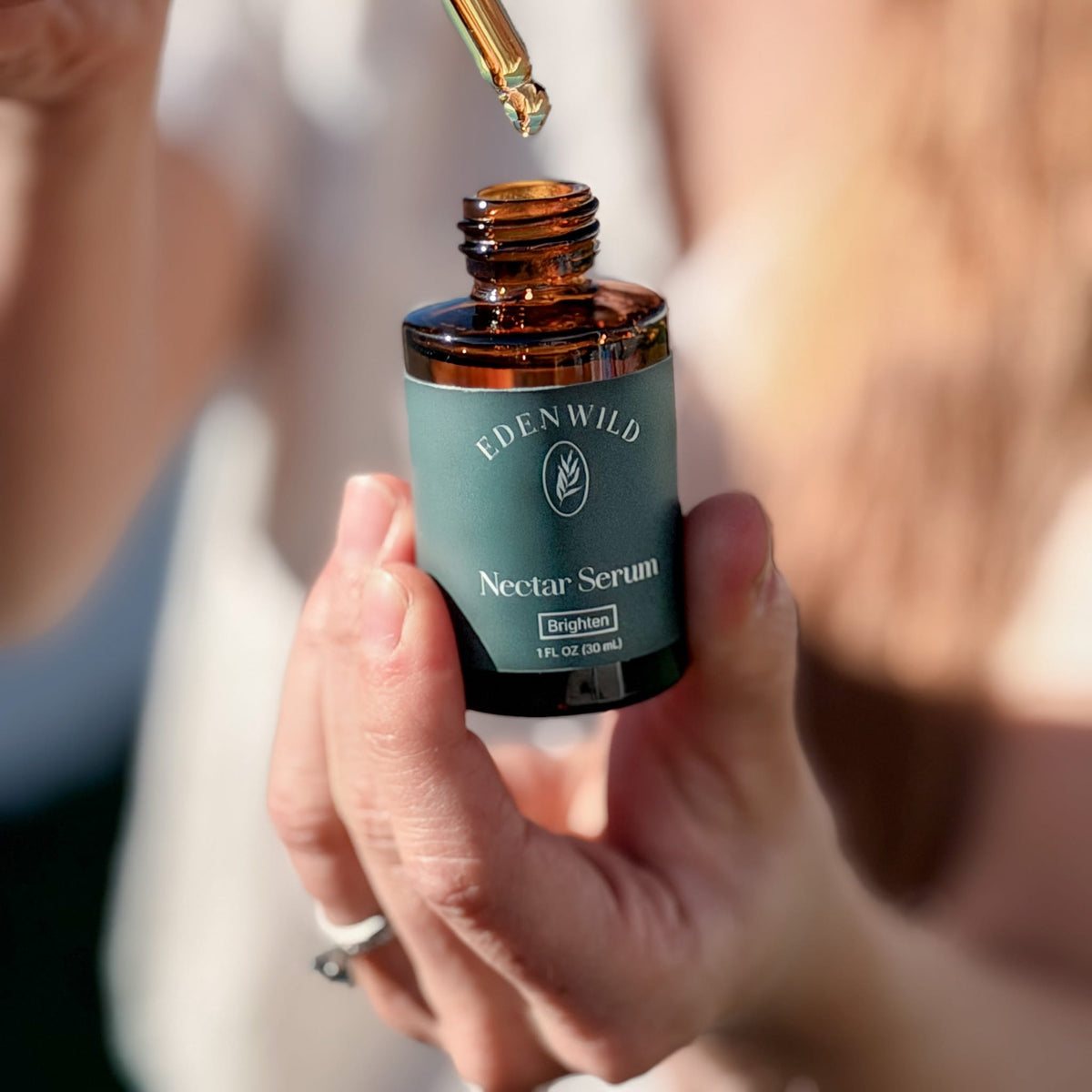 Close-up of a hand holding Edenwild Nectar Serum in an amber glass bottle, with a dropper of golden facial oil above the opening in sunlight.