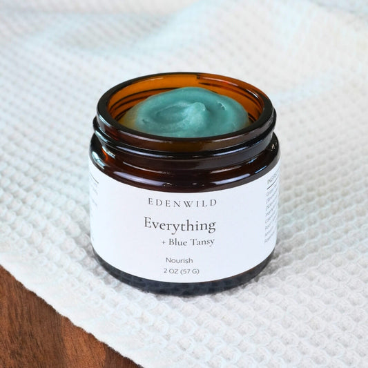 Open jar of grass-fed tallow balm with creamy texture displayed on white towel and dark wood surface, used in Edenwild Everything + Blue Tansy Balm to help calm eczema and sensitive skin