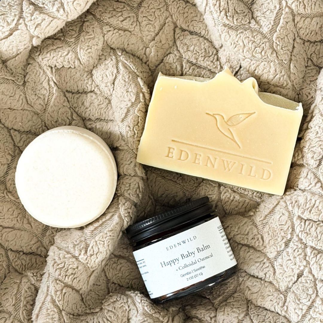 Overhead flat lay of Edenwild Happy Baby Tallow Balm, Bare shampoo bar, and Happy Baby Soap Bar on quilted linen blanket — fragrance-free baby eczema sensitive skincare