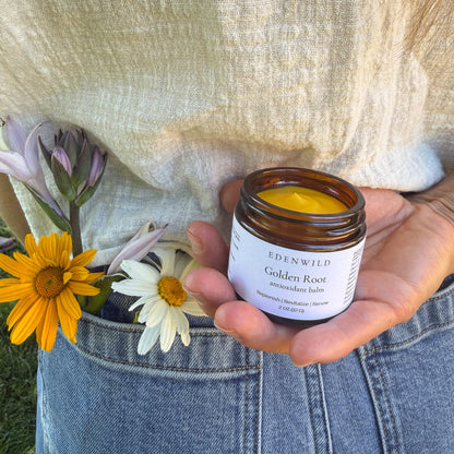 Woman holding Edenwild Golden Root Antioxidant tallow balm outdoors with wildflowers — natural anti-aging moisturizer for aging and sun-damaged skin, 2 oz amber jar