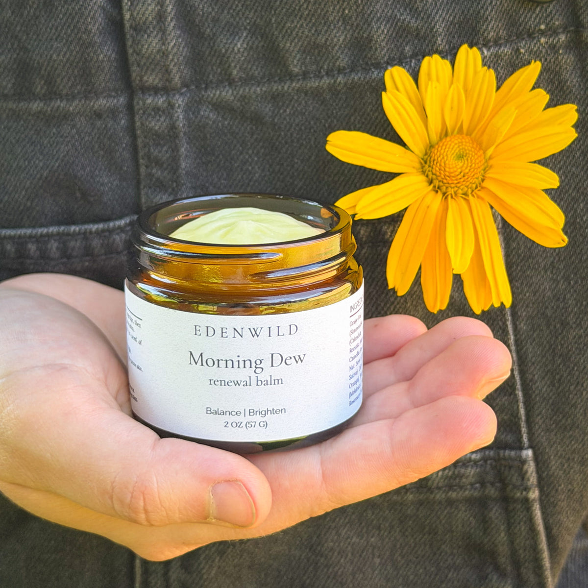 Hand holding open jar of Edenwild Morning Dew Renewal Balm with calendula flower — grass-fed tallow balm for balancing and brightening acne-prone skin