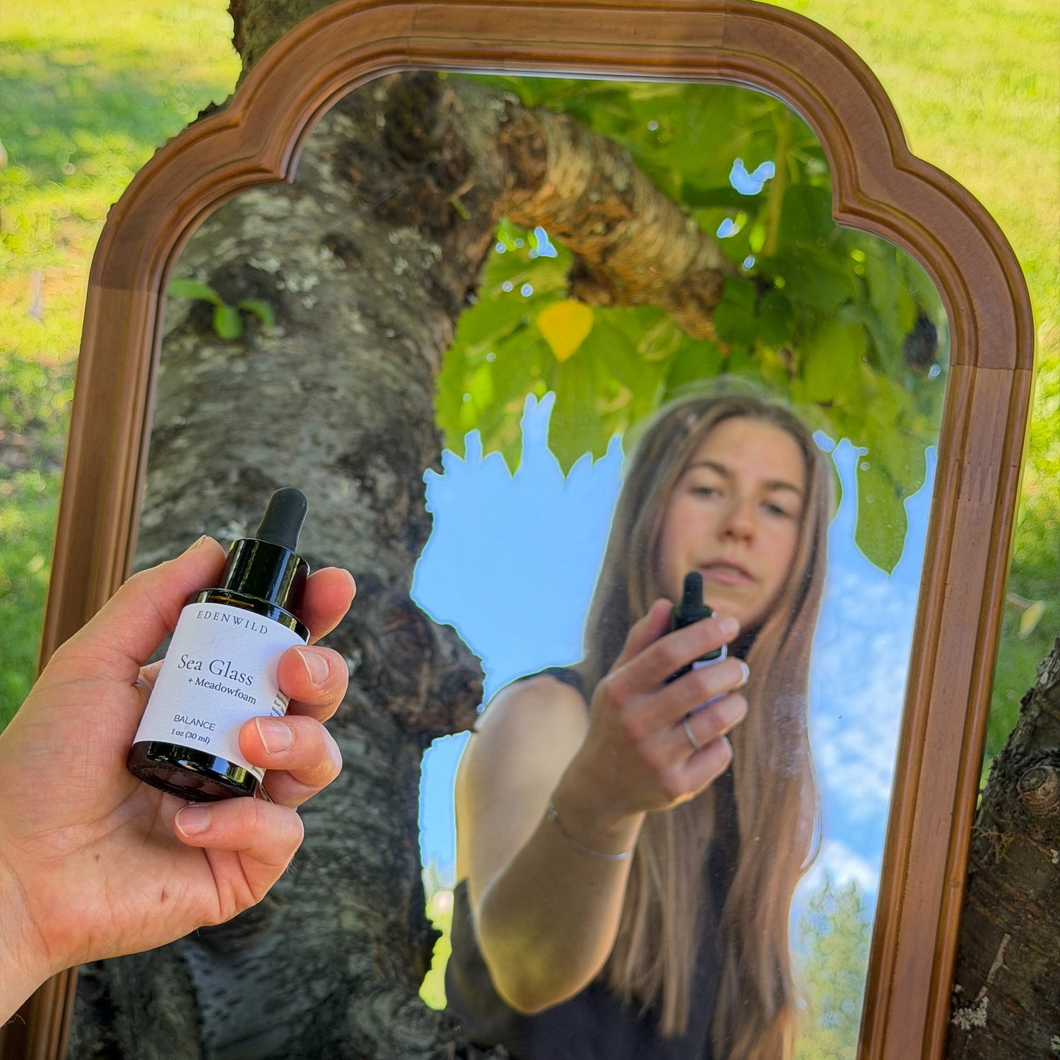 Woman holding Edenwild Sea Glass + Meadowfoam natural face serum outdoors, reflected in a vintage wood-framed mirror — balancing botanical serum for oily skin