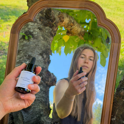 Woman holding Edenwild Sea Glass + Meadowfoam natural face serum outdoors, reflected in a vintage wood-framed mirror — balancing botanical serum for oily skin