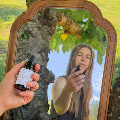 Woman holding Edenwild Sea Glass balancing botanical face serum outdoors with vintage mirror reflection, ancestral skincare